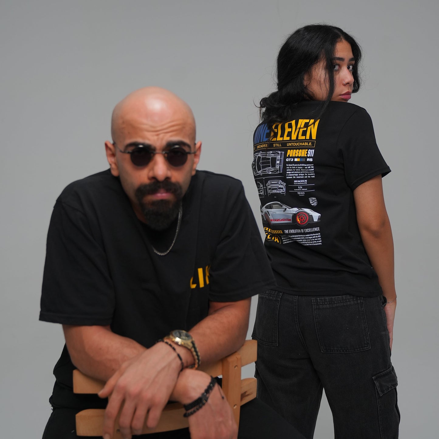 Two people in black shirts with visible text, one sitting on a chair against a gray background.